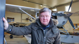 Inne i hangaren finns flera flygplan och väntar på att få komma upp i luften. En man håller upp dörren till en hangar. Bakom honom syns ett litet flygplan.