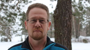 Jesper Sundström säger att det har blivit mera arbete i mörker för brevbärarna, med senare postutdelning och längre arbetsdagar. Jesper Sunström i uteomhusmiljö, snö och och tallar i bakgrunden, i blå fleecejacka