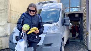 Tuulahannele Halsey har fått mat i kassarna. Hon har också själv frivilligjobbat vid mathjälpen. Ari Vierola i bakgrunden. En glad kvinna tittar ner på ett kålhuvud och en banan som hon har i händerna. Hon står framför en bil som är framför ett hus. I dörröppningen bakom henne syns en man längre bort.