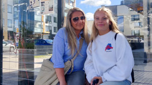 Kristiina Hollsten och Claudia Hollsten väntar på bussen. En lite äldre och en ung kvinna sitter på en bänk i en busskur. Se ler och tittar in i kameran.