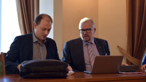 Anders Walls och Ragnar Lundqvist. Två män sitter vid ett bord och ser ner mot några papper. Framför den ena mannen finns en bärbar dator och den andra har en portfölj.