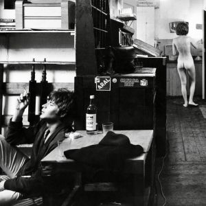 Antti tupakoi pöydän ääressä, alaston Anne seisoo huoneen toisessa päässä elokuvassa Kesyttömät veljekset.