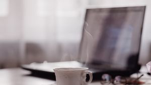 Tietokone ja höyryävä kahvikuppi pöydällä.