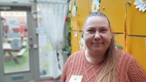 Katri Alifrosti-Holmberg ger på tisdag kanelbullar till sina grannar. Katri Alifrosti-Holmberg, en dam med långt ljust hår i en hästsvans och en rosa tröja, står vid gula skåp.