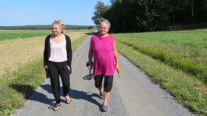 Fanni Sundell och Maria Sundell, här i Tenala. Maria Sundell kommer att fortsätta gå med metalldetektor. "Skatterna" består mest av femcentsslantar och rostiga spikar som hon städar bort från badstränderna. En yngre kvinna, mest klädd i svart och en äldre kvinna klädd i anilinröd t-skjorta och med en metalldetektor i handen går längs en sandväg. Sommar och sol. Åkrar.