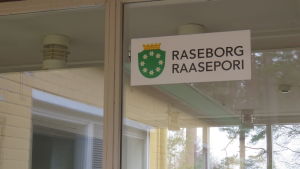 Stället bär ännu spår av sin forna ägare, så här vid den stora ingången. En skylt på en glasdörr med texten raseborg och stadens gröna vapen med krans av vita blommor.