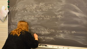 Projektassistens Malin Lundström skriver ner låten. ord, meningar, verser och refräng skrivs på griffeltavlan. En flicka i svart skjorta står med ryggen mot kameran. Skriver "riktiga hästar vill vi ha"