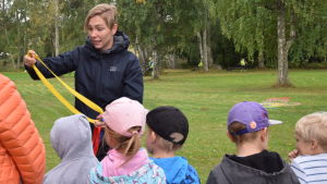 Nu ska det lekas tafatt. Linda berättar reglerna. De är mer avancerade än under redaktörens barndom, men låter riktigt roliga. En kvinna leder en lek med små barn runt sig. De är ute på en grön gräsmatta.