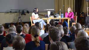 Två kvinnor sitter vid ett bord på en scen. Framför dem sitter en massa högstadie-elever och lyssnar.