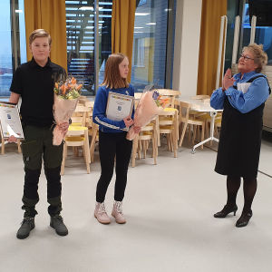 Merja Laaksonen gratulerar Sisu Kymäläinen och Aino Kallio som är duktiga idrottare i Sjundeå. De har diplom och blommor i händerna.