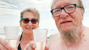 Kuusikymppiset mies ja nainen poseeraavat kameralle ranta-asuissa kuohuviinilaseja kohottaen. Taustalla näkyy meri.