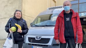 Tuulahannele Halsey och Ari Vierola. En kvinna och en man står bredvid varandra, framför en bil och ett hus. De har kassar i händerna, kvinnan håller också i en banan och ett kålhuvud.