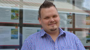 Roland Himmelroos är ny, men ivrig försäljare. En man står ute, framför ett skyltfönster med bilder på hus och bostäder som är till salu.