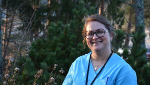 Satu Nurmela säger att tiden får utvisa hur verksamheten vid Karinahemmet tar form. Kvinna i sjukskötarkläder står utomhus och tittar mot kameran. Bakom henne träd och buskar.
