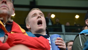 Supportrarna höll liv under hela matchen. En man ropar med stor mun och hejar på sitt lag, Inter Turku under en fotbollsmatch. I handen har han en blå mugg med lagets logo.