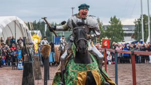 Ritari ratsastaa hevosella turnajaisissa kädessään vasara ja huutaa. Taustalla areena ja yleisöä.
