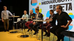 Jarno M. Koponen leder paneldebatt på Yle Betas seminarium för nyhetsrobotik kuusi miestä istuu pöytien ääressä, takaseinällä Yle Beta Day, mediarobotit -juliste