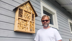 Tomas Järvinen tycker det är roligt att gästerna hittat hit. Att bygga insektshotell är ett sätt att hjälpa pollinerarna som har det allt svårare att klara sig. En man i vit t-skjorta och solglasögon står invid en grå husvägg. På väggen hänger ett insektshotell i trä. Det ser ut som en låda fylld med träklabbar som har hål i sig.