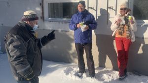 Solveig och Henrik Granholm hann som mest med 30 kaffe med gris-besök ute på Mässkär en vinter. Här i samspråk med Anders Boström från Mässkärsföreningen. En man med ansiktsmask talar med en man och en kvinna som står och dricker kaffe utomhus vid en husvägg.
