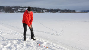 Ulf Eriksson ska slå sitt första slag från bryggan i gästhamnen. En man i röd jacka står uppe på en brygga och ska precis slå iväg en golfboll ut över den vita isen.