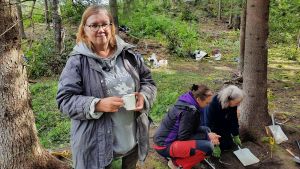 Arkeologen Tuija Kirkinen är ansvarig för projektet. Gruppen har just haft kaffepaus när vi kommer på besök. En kvinna står i en skog med en kaffemugg i handen. Bredvid henne sitter två kvinnor på huk bredvid en grop.