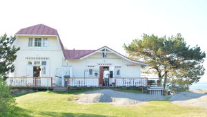 De Fyra Vindarnas Hus finns på Lilla Tallholmen i Hangö och besöks av många turister om somrarna. En gammal vit trävilla med rött tak vid havet, framför dörren står en man.