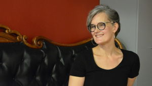 Regissör Tove Appelgren. Kvinna med glasögon sitter på en soffa med en röd vägg bakom sig.