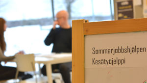 Ingå kommuns ungdomssektor ordnar sommarjobbshjälpen två gånger i år. En skylt där det står Sommarjobbshjälpen. I bakgrunden skymtar två personer vid ett bord.