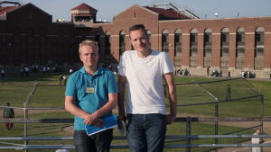 Programmakarna Tomas Lindh och John Stark vid Atticafängelset i USA. Två män står framför en tegelbyggnad med en sportplan i bakgrunden och metallstaket runt.