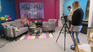 Kårkullas tv-studio. Två personer filmar en tredje person som sitter i en soffa i ett färggrant rum.