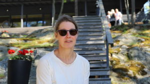 Irma Puttonen. En kvinna i solglasögon sitter på en trappa vid en stuga utomhus.