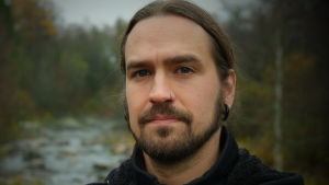 Mathias Lillmåns sjunger i Finntroll.. Mathias Lillmåns ser in i kameran, i bakgrunden skog och en fors. Svart jacka. Långt hår i svans. Piercing i näsan.