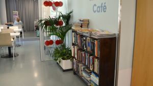I det rum i aulan som kallas Vardagsrummet finns bland annat en bokhylla. Bokhylla och grönväxter, i bakgrudnen cafébord.