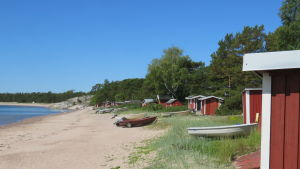 Olika sorts sidobyggnader kan behövas om många delar samma ställe under sommaren. Arkivbild. Hangö, Gunnarstrand en solig sommardag med knallblå himmel. Rödmålade fiskebodar och båtar längs sandstranden.
