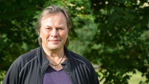 Jan Jacke Jacobson. porträttbild av en man. Han står ute i grönskan och bär en svart jacka över en lila t-skjorta.