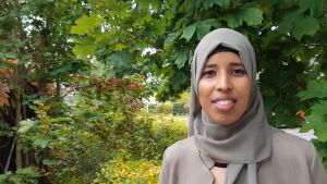 Samira Abdullah bor just nu bara på veckosluten i Borgå. Hon bor i veckan hos en kompis i Helsingfors för att ha kortare pendlingsväg. I februari i nästa år tänker hon flytta till Esbo. En kvinna med en grå dräkt och huvudduk ler och tittar in i kameran. I bakgrunden gröna löv med inslag av höstfärger.