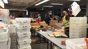 Det råder organiserat kaos innan de behövande släpps in till Hurstis mathjälp. En man lyfter en låda, andra packar ur lådor.