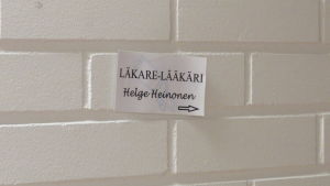 En liten anspråkslös lapp på väggen visar var Helge Heinonen har sin läkarmottagning. En liten papperslapp på en vit tegelvägg (inomhus). Det står Läkare-Lääkäri Helge heinonen på lappen.