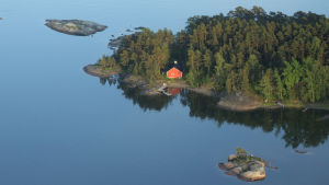 En ö med en röd stuga och blått vatten omkring i Ingå skärgård.