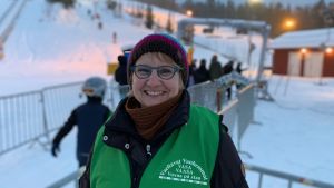 Elisabeth Henriksson klär sig gärna i den gröna västen och ställer upp för att bidra till större trivsel vid Öjberget. Fler föräldrar får gärna komma med i frivilligverksamheten säger hon. Leende kvinna utomhus. I bakgrunden köar slalomåkare till skidlift. Kvinnan är klädd i grön väst där det står Vasa - vuxna på stan.
