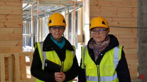 Forsby skolas rektor Helena Järventaus och Koskenkylän koulus rektor Katja Smeds ser fram emot att få flytta in Två kvinnor i skyddsväst och hjälm poserar framför en stockvägg