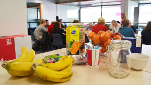 Mellanmål för att hålla kreativiteten vid liv. På ett bord står frukt, saft, en tom glasburk och små chokladpåskägg i en påse. Bakom matvarorna syns flera människor som sitter runt ett långt bord.