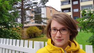 Mariann Hilli är lärare för den svenska utbildningen. Kursdeltagarna kommer från hela Nyland. En kvinna i gul jacka med halvlångt brunt hår och glasögon står vid ett vitt staket, i bakgrunden finns våningshus och en grön gräsmatta.