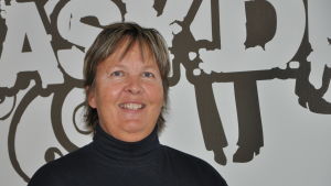 Ulla Nummelin. Närbild på en leende Ulla Nummelin som står framför en vägg med bilskolans logotyp i bakgrunden.