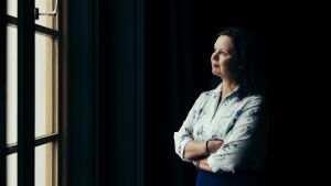 Teaterchefen Ann-Luise Bertell vid Wasa Teater. På bilden ses författaren Ann-Luise Bertell titta ut genom ett fönster iklädd en ljus skjorta och blå kjol mot en mörk bakgrund.