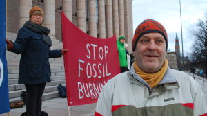 "Det är en paradox att världen förfasas över coronaviruset, fast klimathotet kan ge mycket större konsekvenser för mänkligheten", säger Juhani Lehto. Klimatdemonstrant på riksdagens trappa, med röd banderoll med texten "Stop fossil burning".