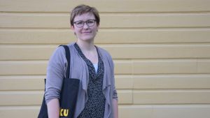 Janiina Mieronkoski är handledare på Brottsofferjourens serviceställe i västra Nyland. En kvinna med glasögon står framför en gul vägg, hon har en tygkass med gul text på, det står Riku på den.
