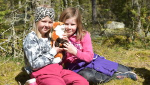 Hanna och Tyra är redo för att bygga. Hanna och Tyra sitter i gräset med skogen som bakgrund. De håller i ett mjukisdjur som föreställer en räv.