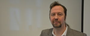 Mikael Mäki. Mikael Mäki, en man med mörk mustasch, skägg, hår i hästsvans och en brun kavaj.