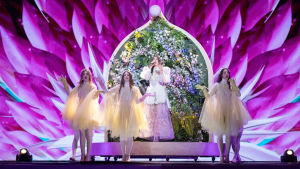 Greklands Katerine Duska i Eurovisionen 2019. Greklands Katerine Duska framför en blomstervägg med tre vitklädda dansare.